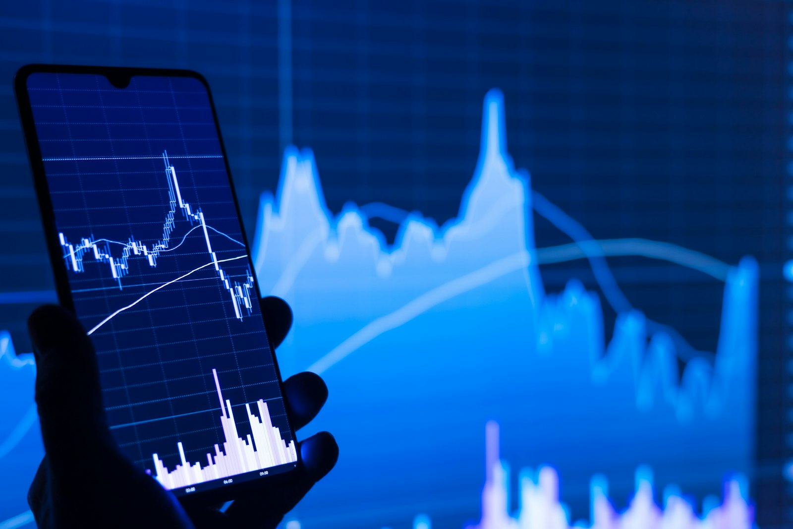 Silhouette of a hand holding a phone showing a stock price chart against a blue trading screen